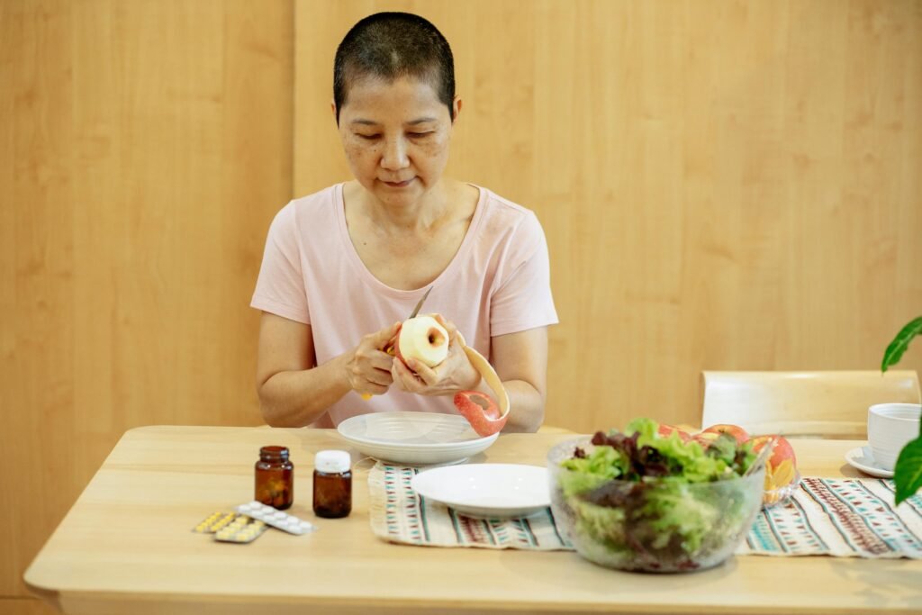 Middle aged ethnic female with short hair in t shirt peeling fresh apple and preparing salad while sitting at table with veggies and various medicine during remission