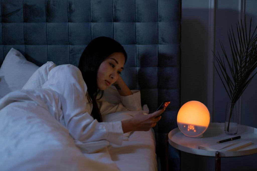 Asian woman in bed using smartphone, illuminated by warm light, indicating insomnia or sleeplessness.